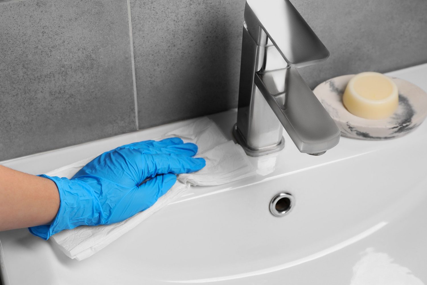Hand mit blauem Handschuh wischt ein weißes Waschbecken neben der Armatur ab.