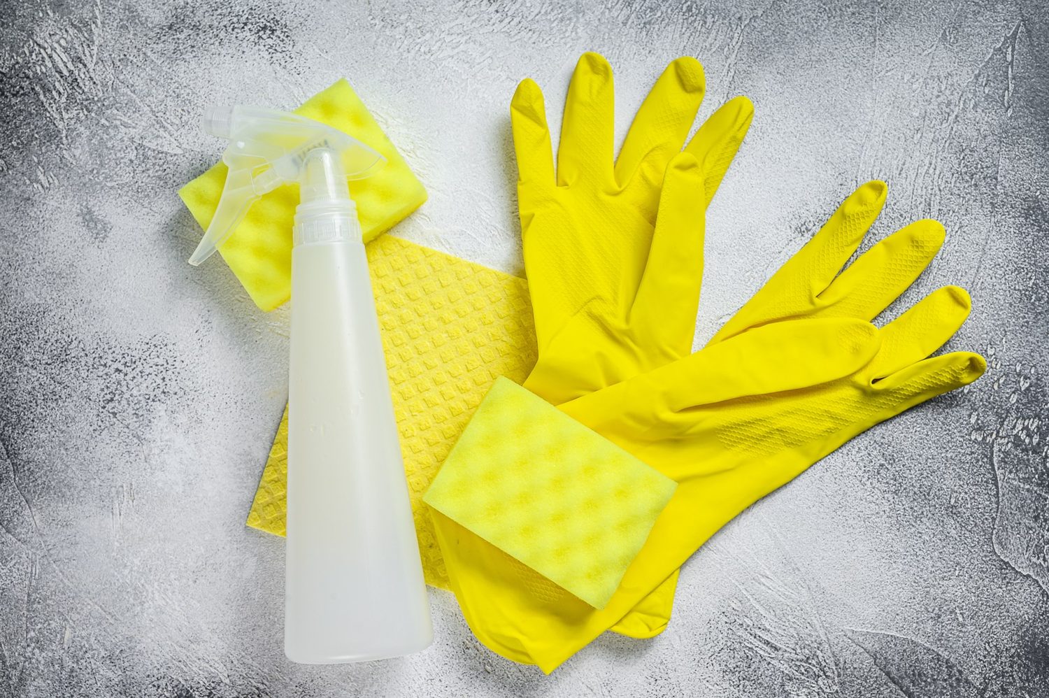 Gelbe Gummihandschuhe, zwei gelbe Schwämme, ein gelbes Tuch und eine weiße Sprühflasche liegen auf grauer Fläche.