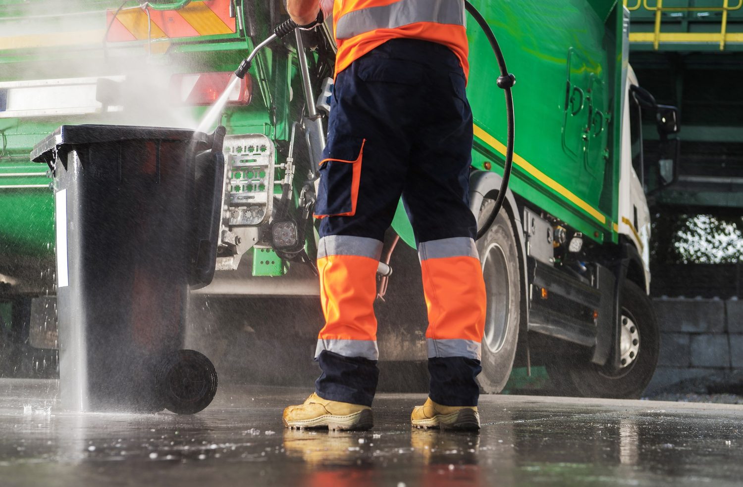 Mitarbeiter reinigt eine Mülltonne mit einem Hochdruck-Wasserstrahl neben einem Entsorgungsfahrzeug