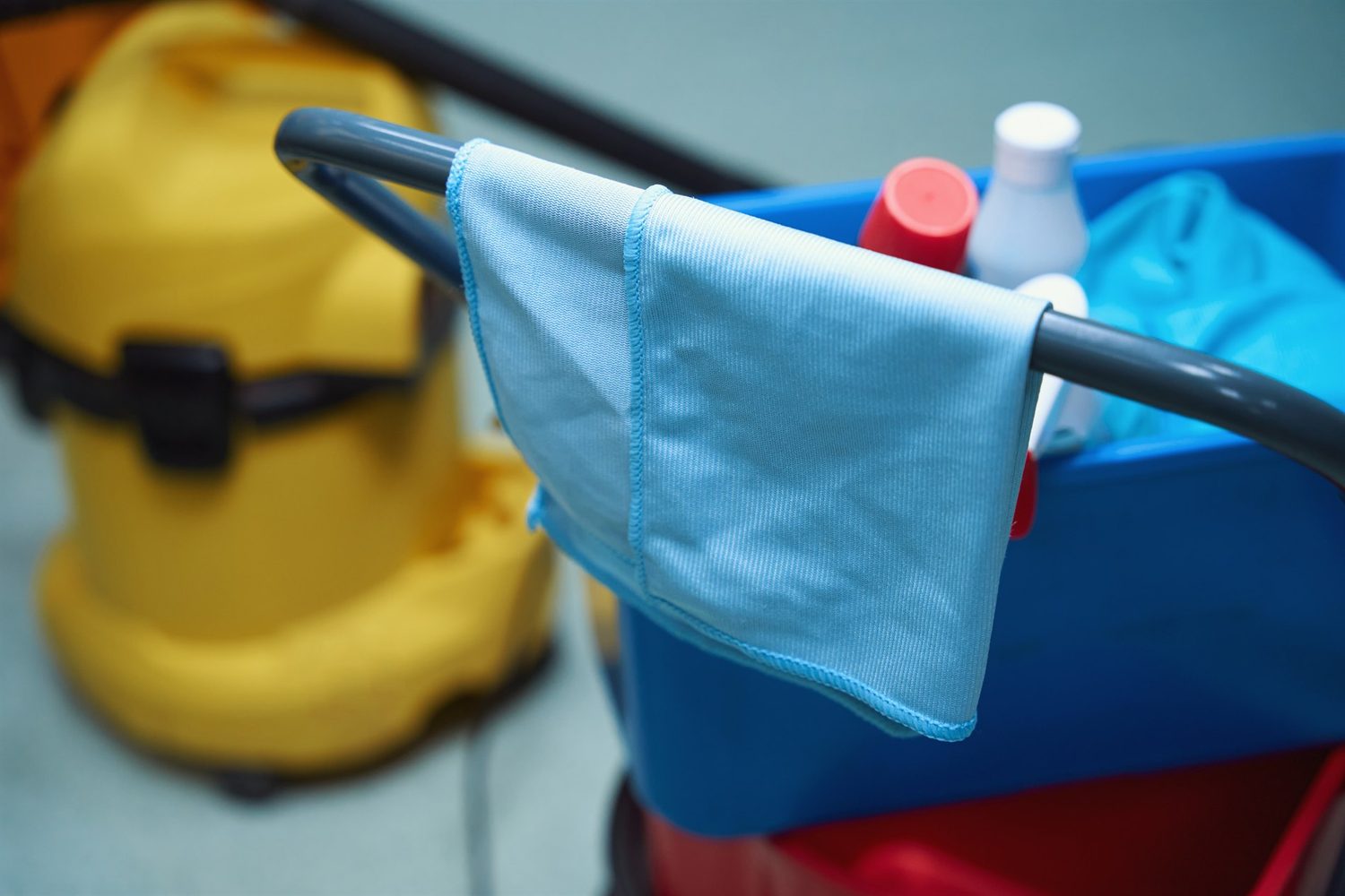Ein blaues Reinigungstuch hängt über dem Griff eines Materialwagens mit Flaschen; ein gelber Staubsauger steht unscharf im Hintergrund.