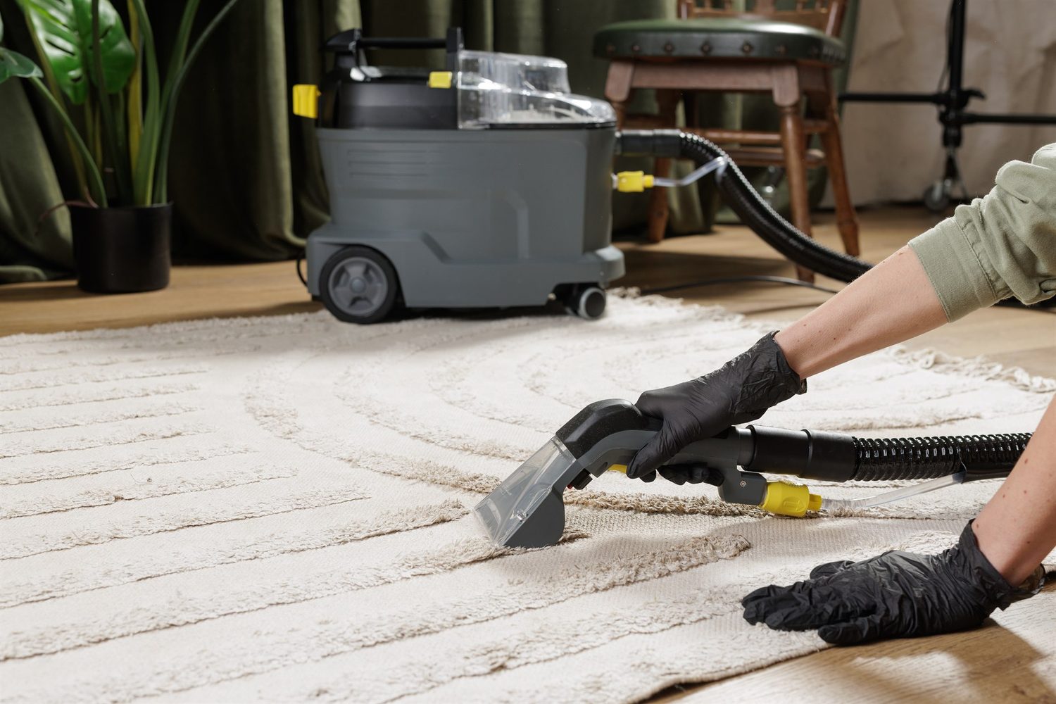 Eine Person mit schwarzen Handschuhen führt die Extraktionsdüse eines Waschsaugers über einen hellen Teppich.