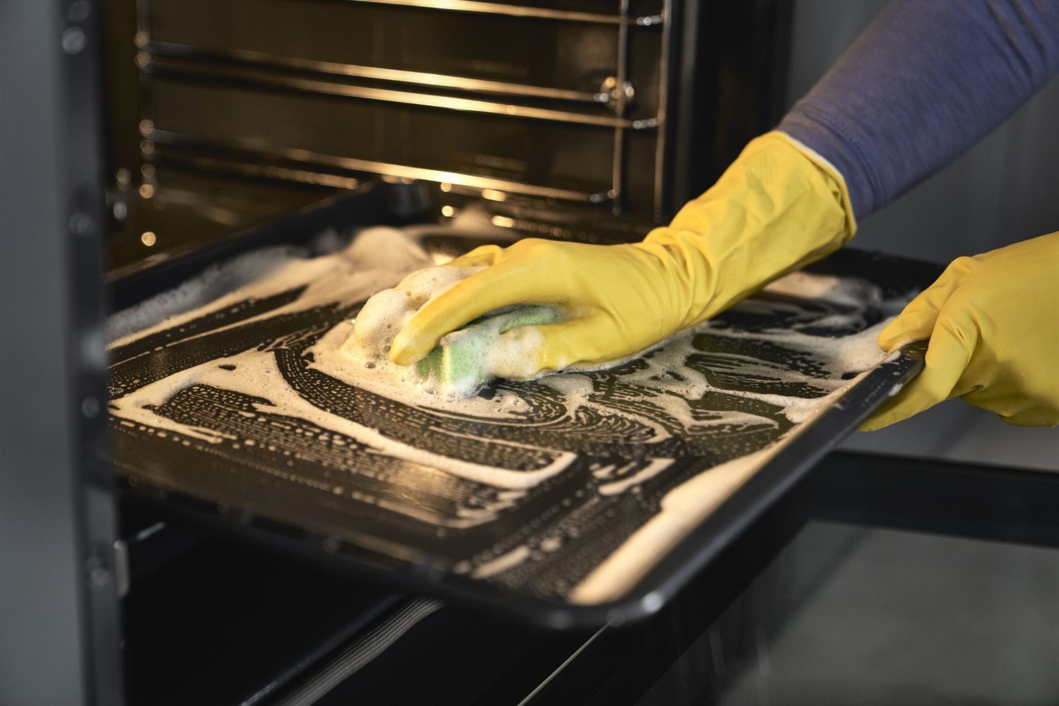 Hände mit gelben Gummihandschuhen reinigen ein verschmutztes Backblech mit einem Schwamm und Schaum vor dem geöffneten Backofen.